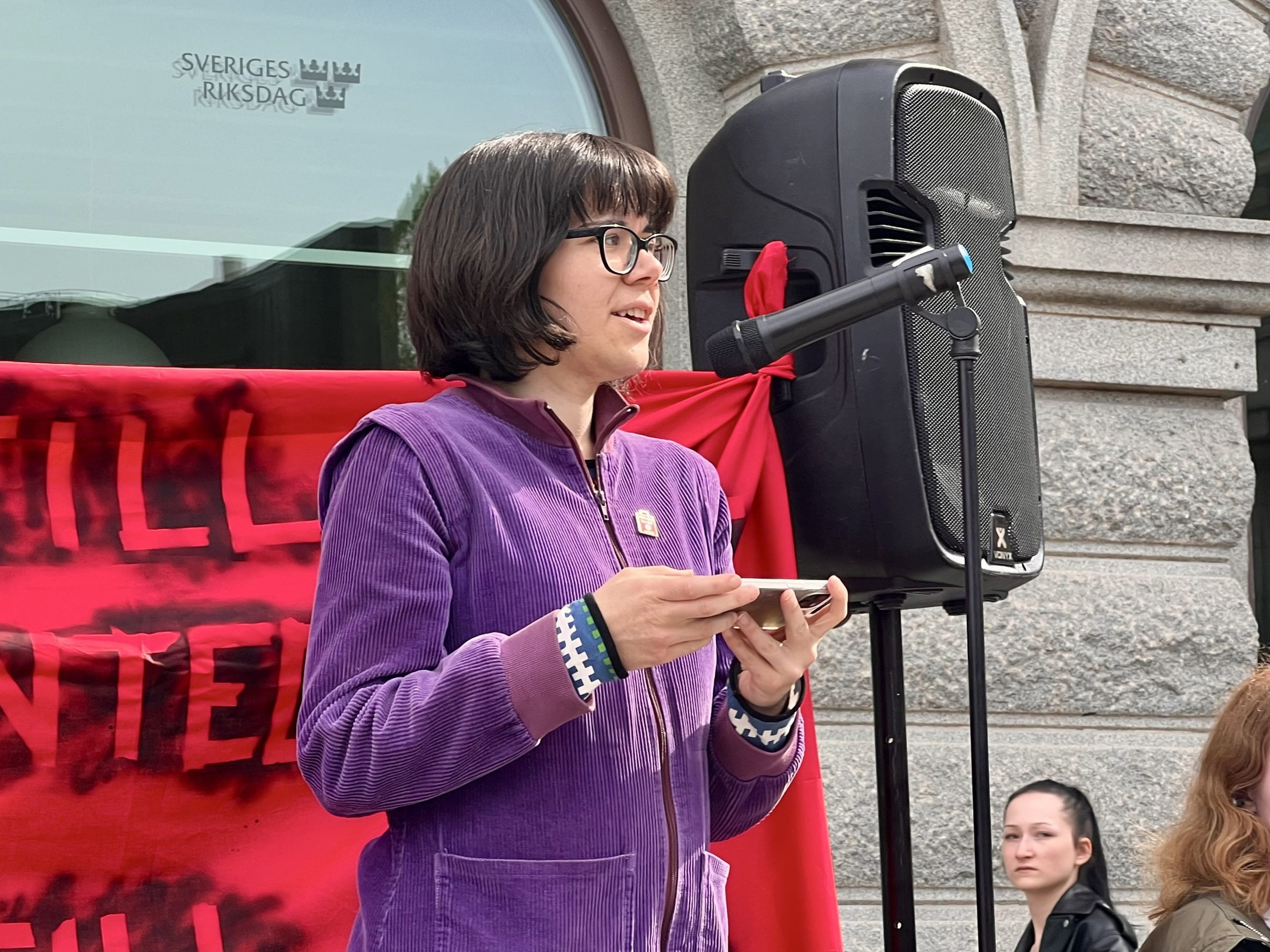 Andrea Andersson-Tay, riksdagsledamot för Vänsterpartiet, talar vid demonstration. En stor högtalare syns i bakgrunden.