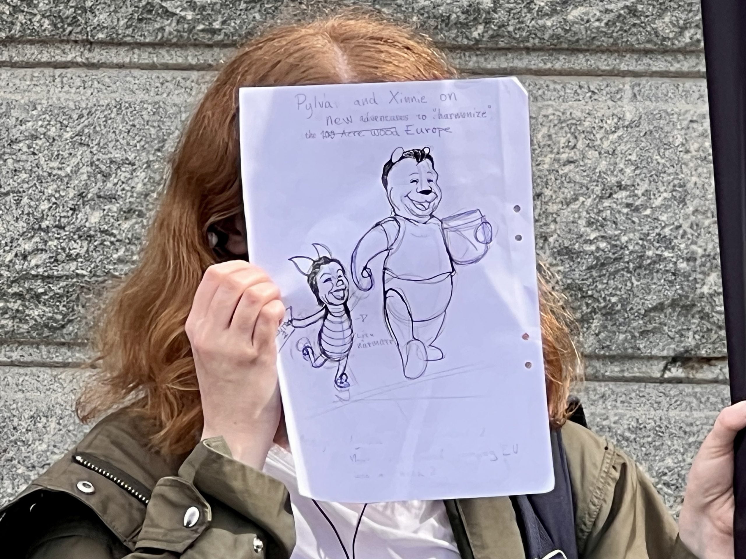 Okänd person håller upp en satirteckning med texten ”Pylva and Xinnie on new adventures to “harmonize” Europe” (på svenska: Pylva och Xinnie på nya äventyr för att ”harmonisera” Europa). Texten ”Europe” (på svenska: Europa) ersätter överstrukna ”100 acre wood” (på svenska: Hundramilaskogen).
