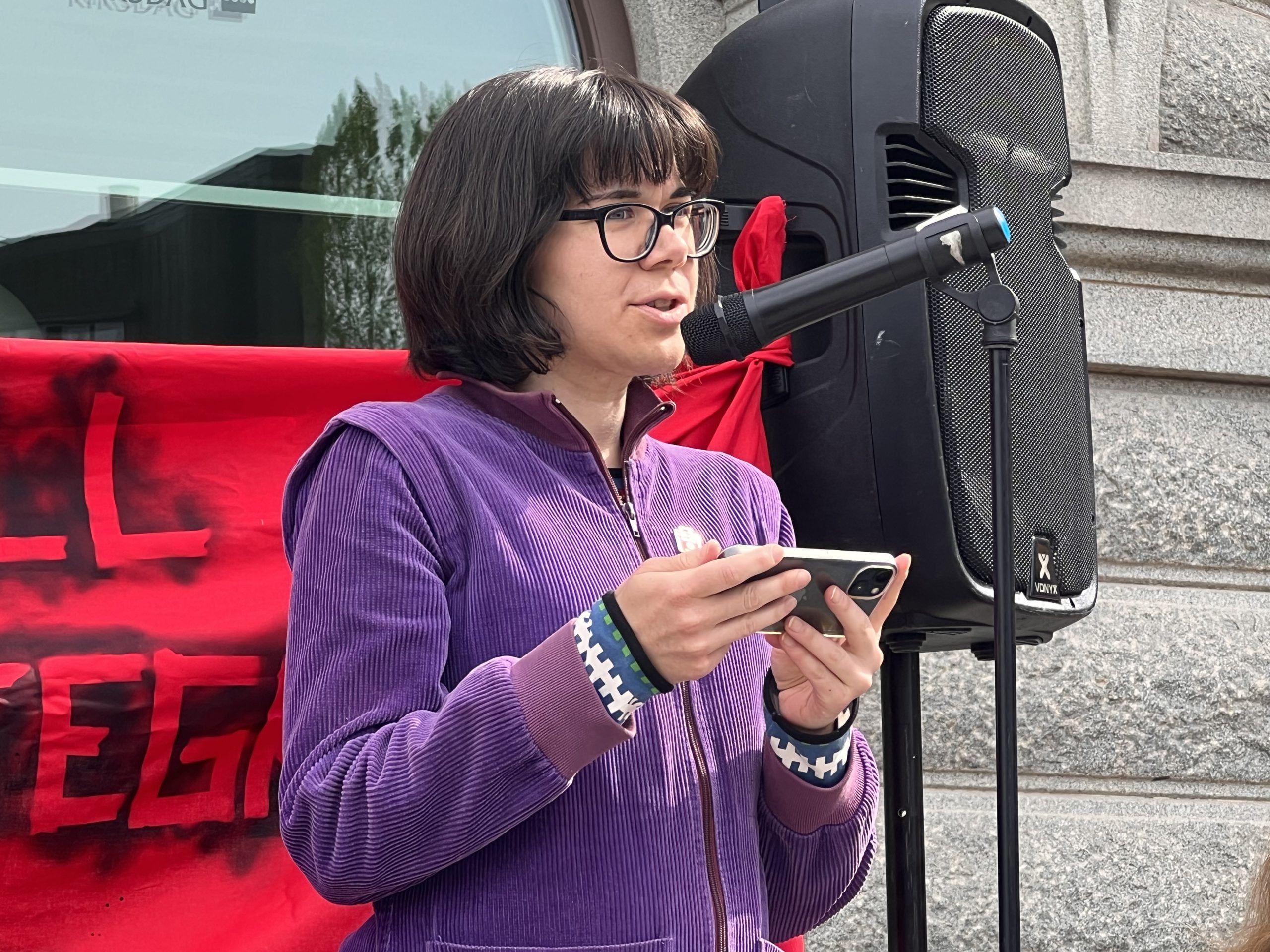 Andrea Andersson-Tay, riksdagsledamot för Vänsterpartiet, talar vid demonstration. En stor högtalare syns i bakgrunden.