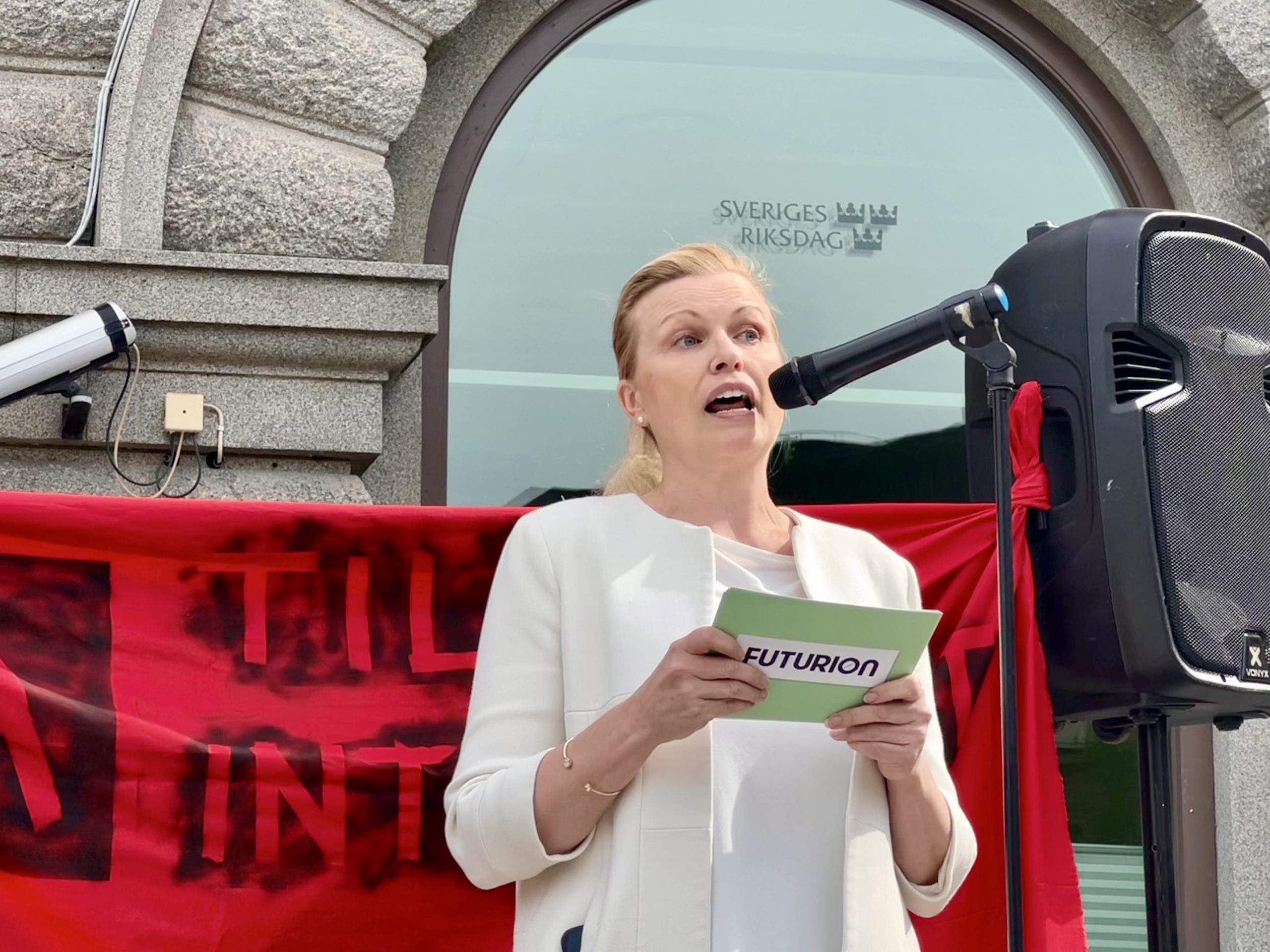 Ann-Therese Enarsson (vd på tankesmedjan Futurion) talar vid demonstration. Ett fönster med Sveriges Riksdags logotyp syns i bakgrunden.